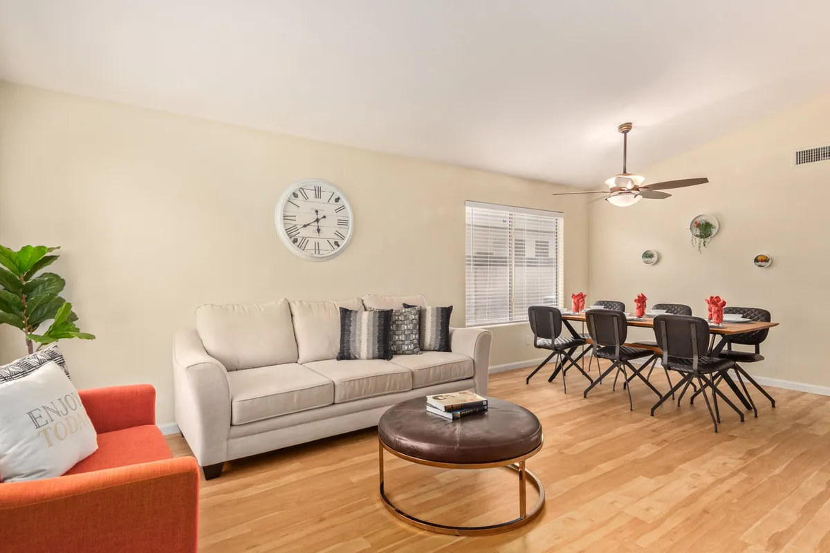 The image shows a cozy interior featuring a comfortable sofa, a dining table that could double as a game table, a large wall clock, and a decorative plant. The open arrangement makes the space suitable for both relaxation and interactive activities like board games or casual meetings.