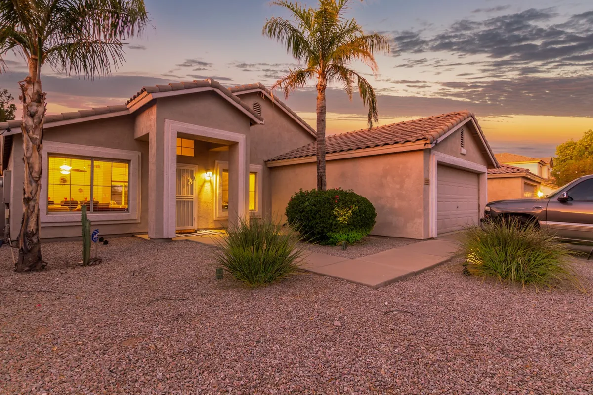 A welcoming front porch framed by palm trees and a landscaped garden, with comfortable seating that invites guests to relax or enjoy a morning coffee. The garage and tidy exterior also hint at convenience and easy access.