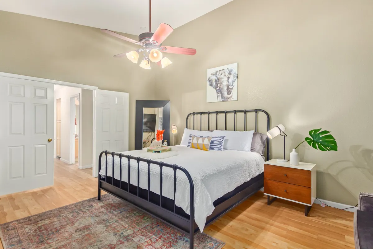 A serene bedroom with a cozy bed, nightstand, elegant wall art, a ceiling fan, and a soft area rug. The space is bathed in natural light, creating a peaceful and restful atmosphere ideal for relaxation or reading.