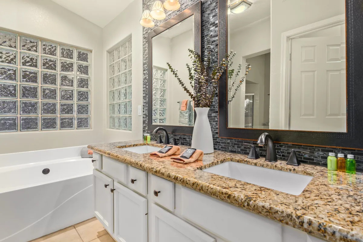 A stylish en-suite bathroom with a double sink vanity featuring a granite countertop, large mirror, glass block wall providing privacy and light, and a spacious bathtub. The room feels modern, tranquil, and perfect for unwinding.