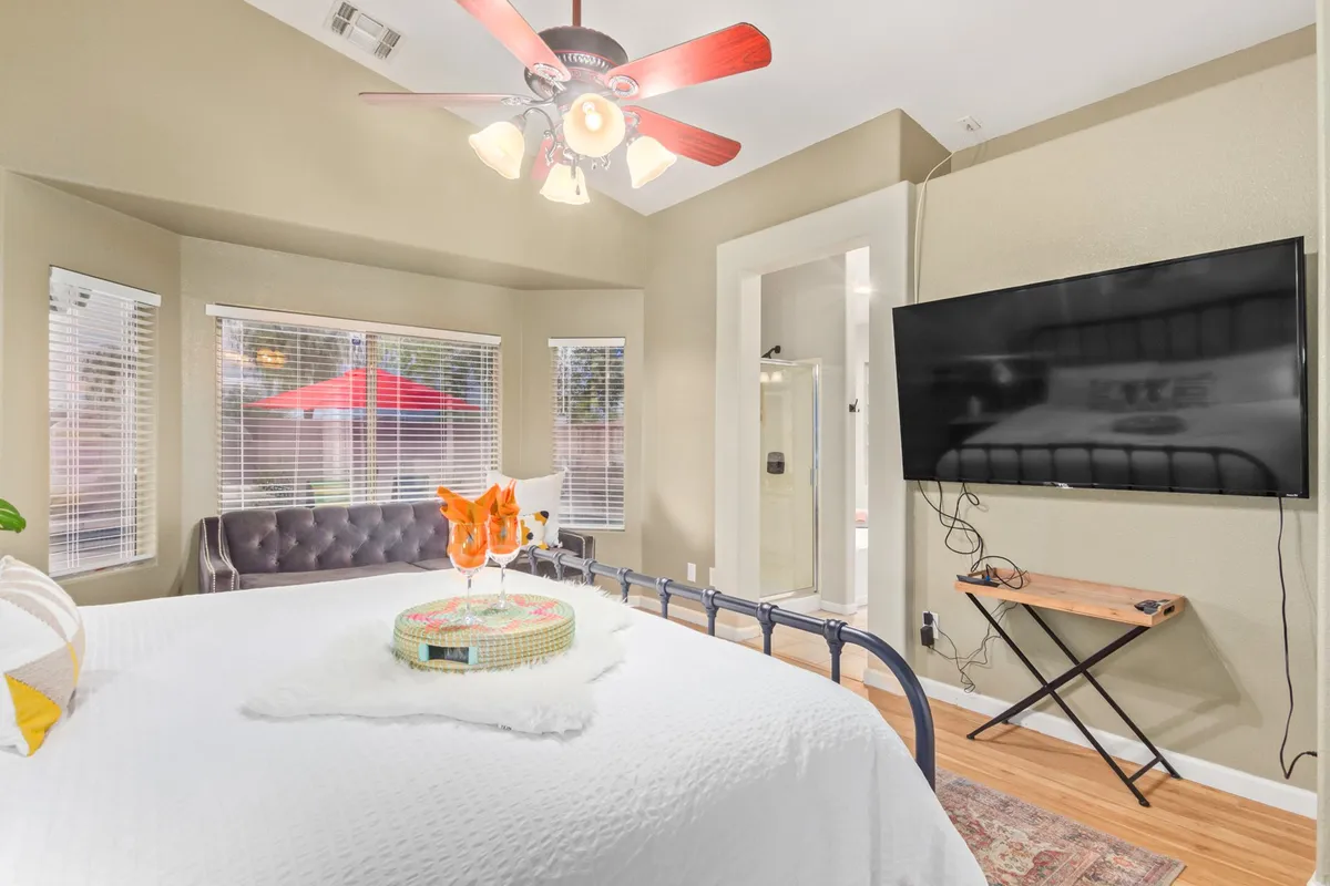 A spacious bedroom featuring a king-sized bed with decorative pillows, a small table with drinks, a large TV mounted on the wall, a comfortable couch in the corner, and bright windows with blinds that let in natural light. The room exudes comfort and is thoughtfully arranged for relaxation and privacy.