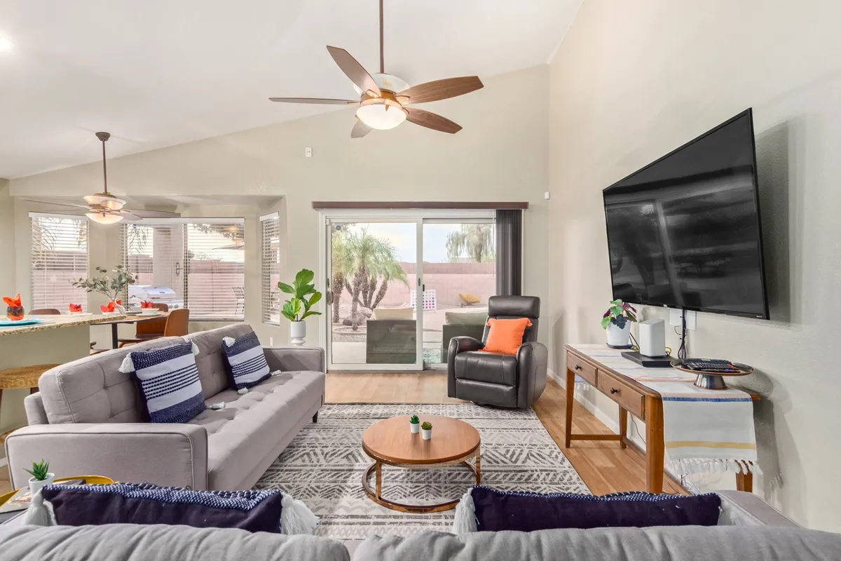 This image showcases a spacious living area with a large, comfortable sofa, a recliner, a super-sized TV, a central coffee table, and a bright window view. The natural light fills the space, emphasizing the room's warmth and openness.