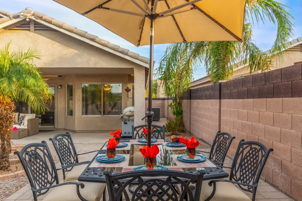 A cheerful backyard setting with a dining table adorned with red flowers, an umbrella for shade, swaying palm trees, and the home in the background. This inviting outdoor space is perfect for gatherings or relaxed moments enjoying the sun and fresh air with your dog.