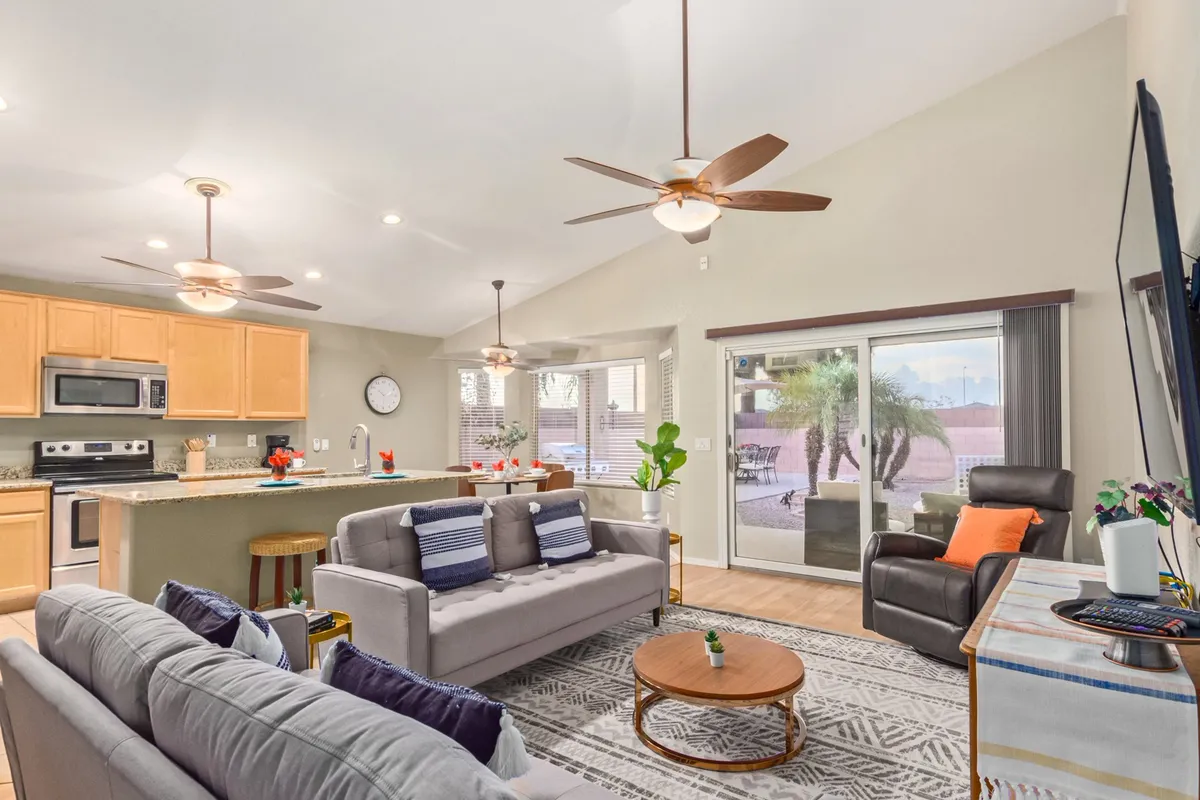 A wide-angle view of a combined living and dining area, featuring multiple sofas, a kitchen island, a large window letting in sunlight, ceiling fans, and a dining table ready for family meals or game nights.