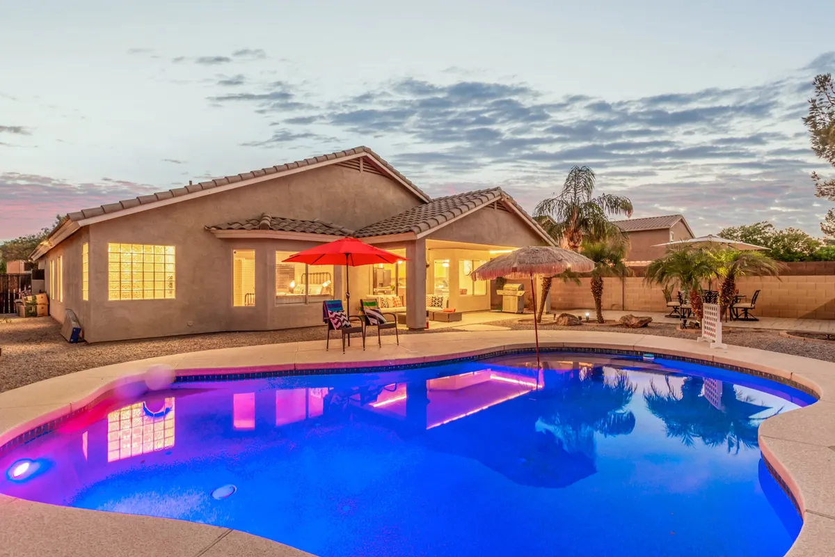 A sparkling swimming pool is the centerpiece of the backyard, surrounded by red umbrellas, patio seating, and the house in the background. The space exudes a tranquil yet festive atmosphere, perfect for swimming, sunbathing, or relaxing outdoors.
